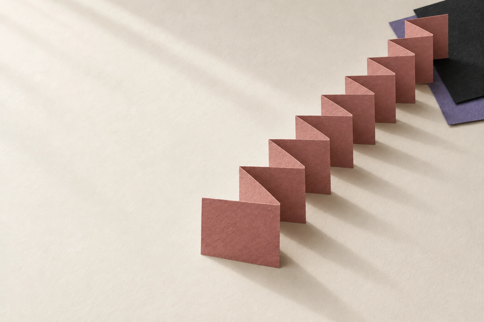 A long rose-colored paper strip folded into twelve panels stands on warm white paper, with charcoal and muted violet sheets weighing down one end.