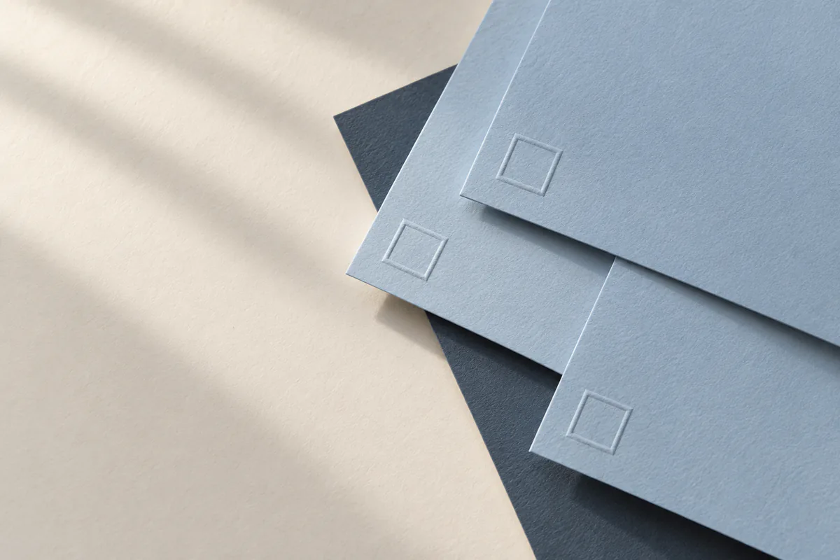 Drei hellblaue Papierbögen liegen übereinander. In jeder Ecke befindet sich ein leerer quadratischer Prägedruck. Darunter ist dunkelgraublaues Papier zu sehen.