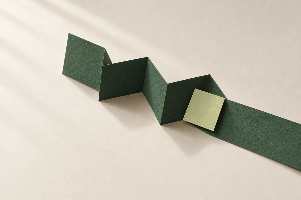 Ein langer dunkelgrüner Papierstreifen ist zu Seiten gefaltet. Die zweite Hälfte liegt flach, an einer Falte liegt ein kleines hellgrünes Papierstück.